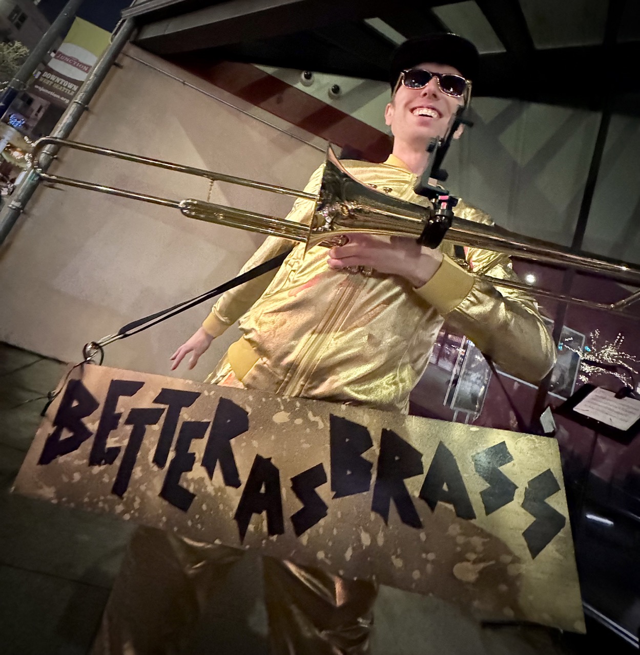 Man wearing metallic gold clothes and dark sunglasses holding a trombone with a sign hung around him reading Better As Brass